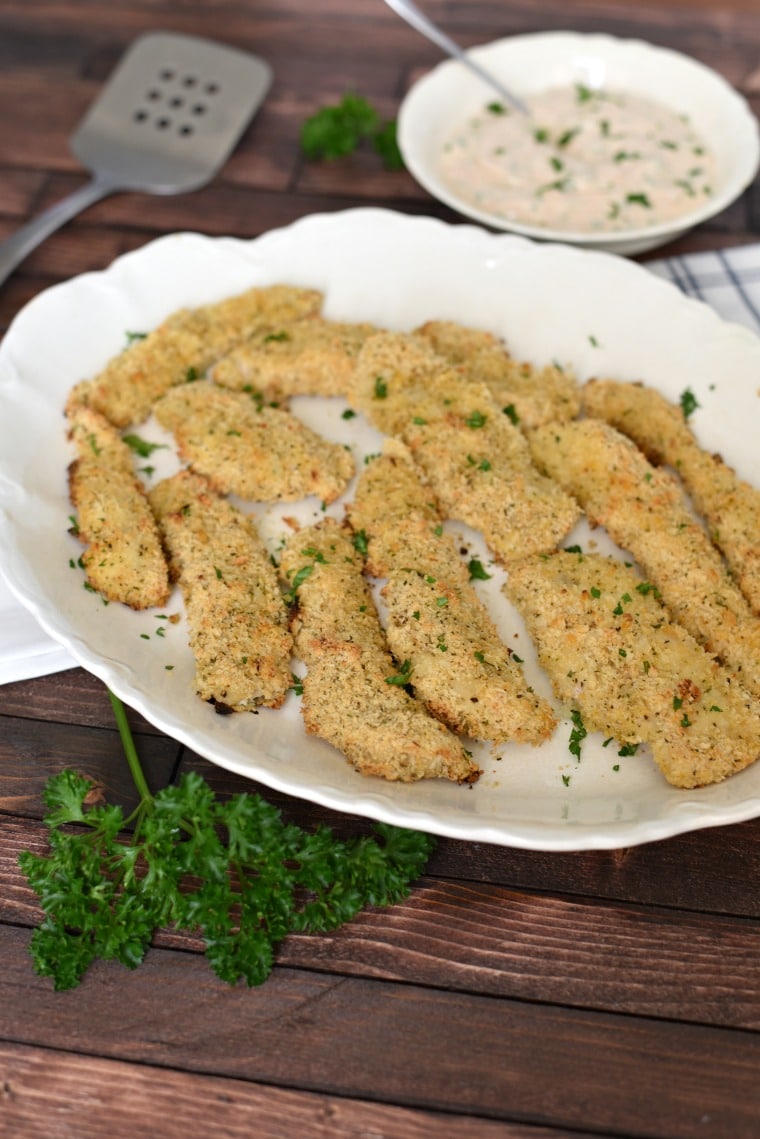 A white platter with panko breaded chicken strips on a wooden table with parsley 