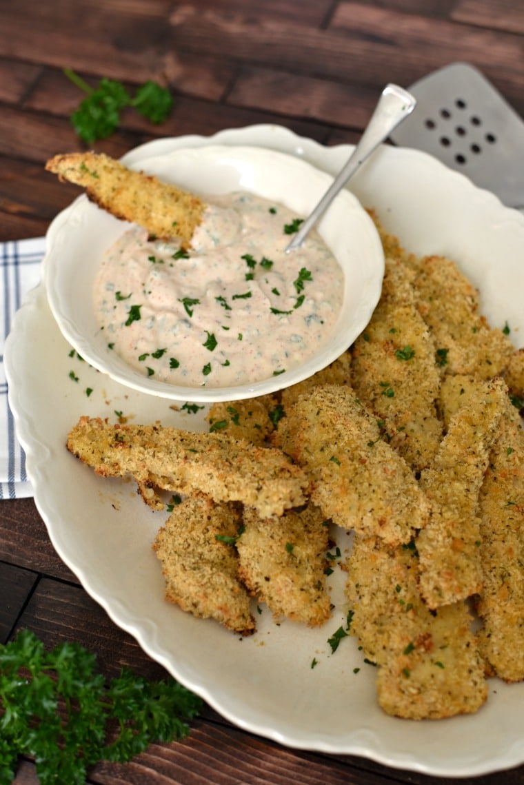 A platter of panko breaded chicken strips with one strip dipped in a white bowl of creole dip 