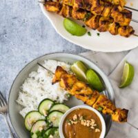 plate with rice, cucumber salad, chicken on skewers and bowl of peanut sauce next to tray with chicken skewers