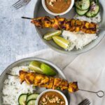 two plates with chicken satay and peanut sauce, rice and cucumbers