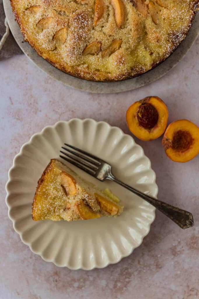 German peach kuchen slice on white plate with fork beside platter of more cake and sliced peach