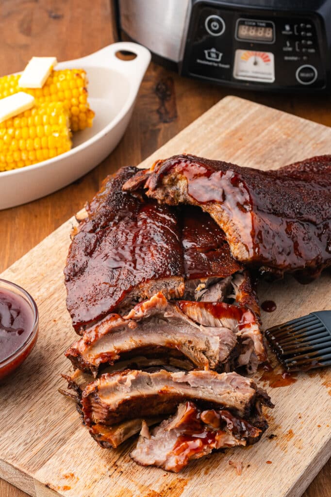 slow cooker baby back ribs on wooden cutting board with slow cooker and dish of corn in background