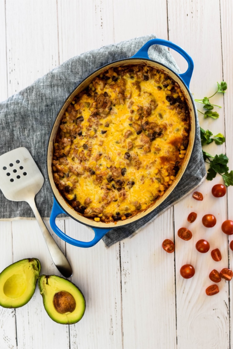 baking dish with beef enchilada casserole, surrounded by serving spatula, tomatoes, and avocado slices