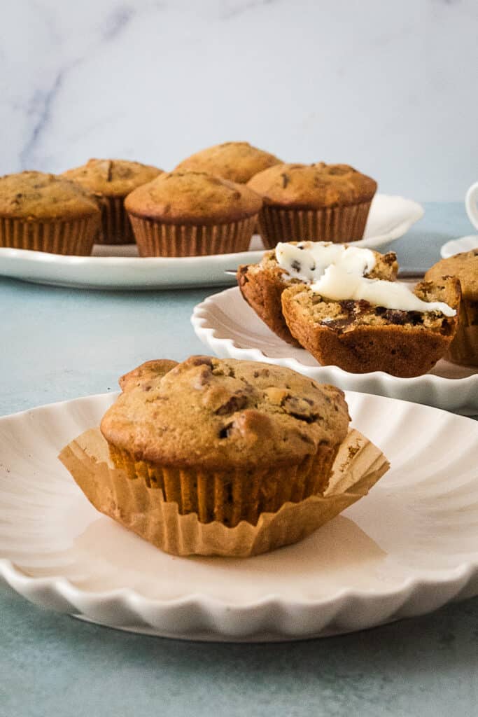 closeup of muffin in a muffin liner on a white plate, with second plate of muffins in background and platter with more muffins behind that