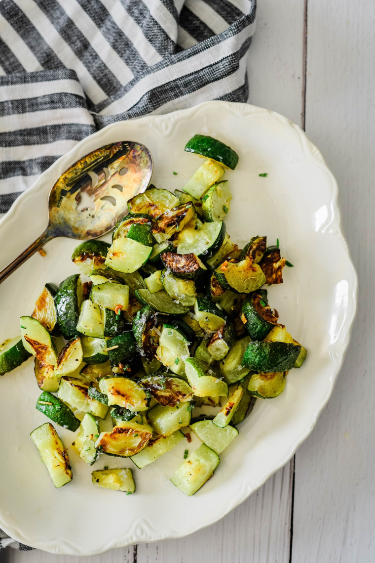 roasted zucchini on white serving platter with antique spoon beside, black and white striped towel behind