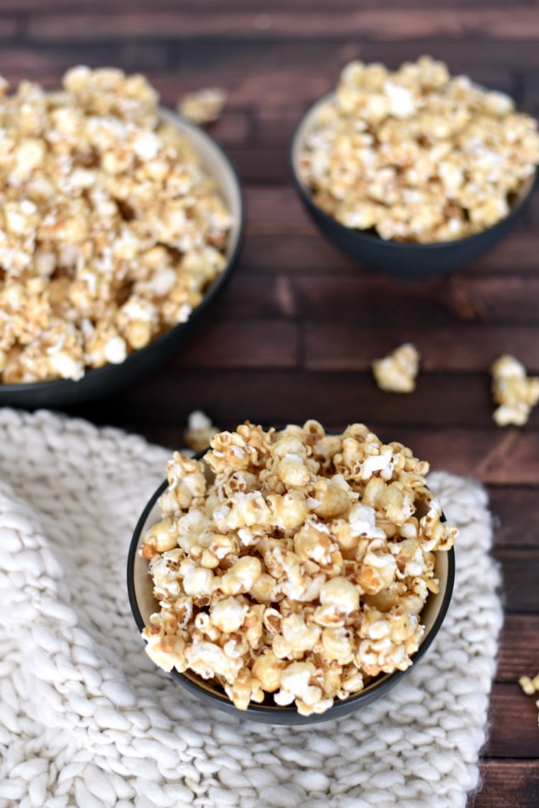 Three bowls of peanut butter popcorn sitting on a white knitted blanket, which is sitting on a wooden table