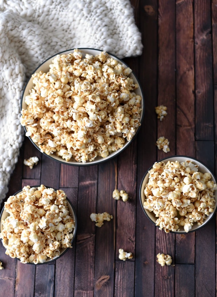 Three bowls of peanut butter popcorn sitting on a wooden table. Some popcorn kernals are spilled around the table and a white towel is on the table too