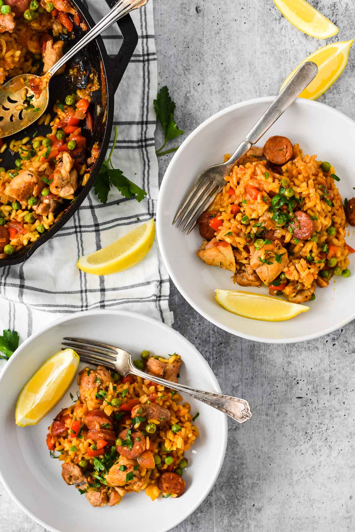 two bowls with paella next to cast iron skillet, lemon slices and forks arranged around