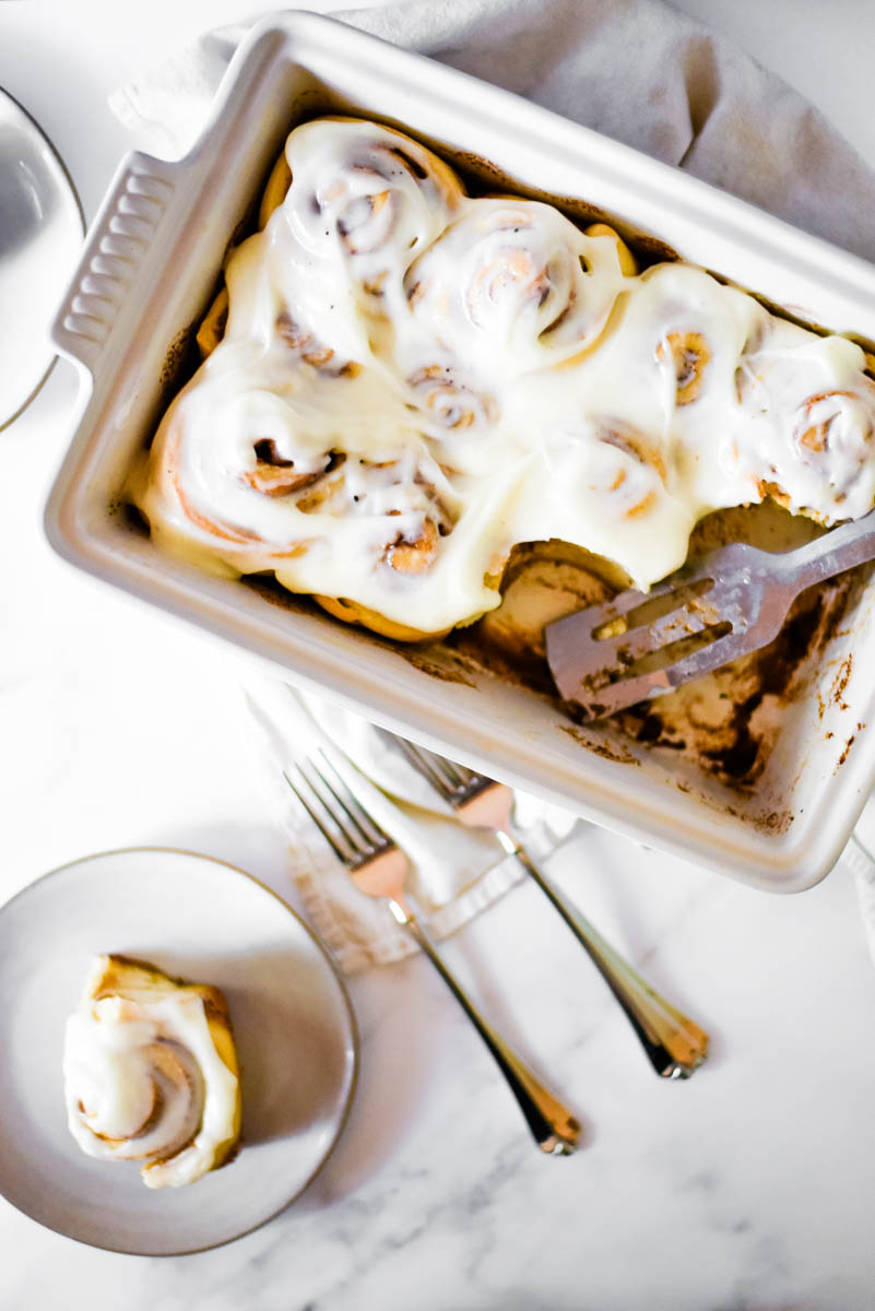 overhead view of white baking dish with sourdough cinnamon rolls, serving utensil on the side and white plate and two forks beside