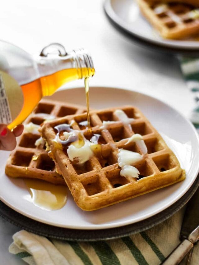 girls hand pouring maple syrup onto two sourdough waffles on white plate with silverware beside