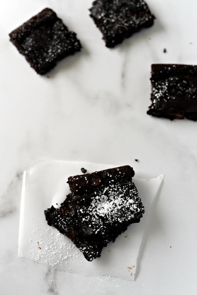 Two stacked black bean brownies dusted with powdered sugar sitting on a napkin on a white marble countertop. Two other brownies on the countertop