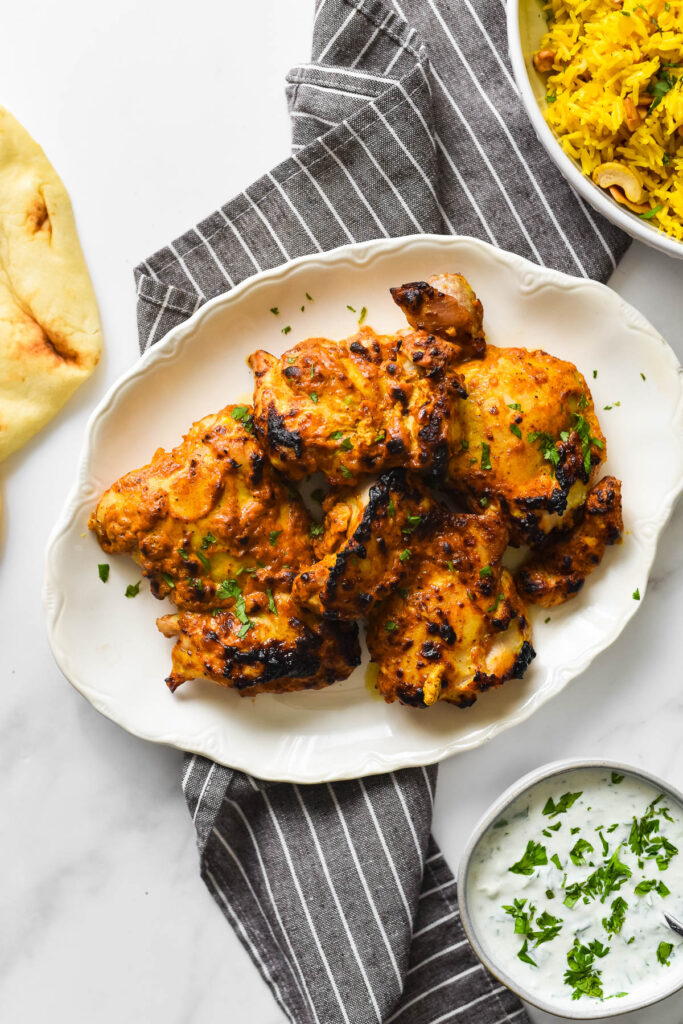 Grilled chicken thighs on platter surrounded by naan, yellow rice, and raita dipping sauce