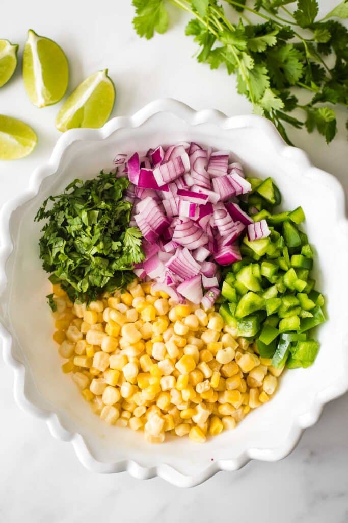 white bowl with corn, chopped peppers, chopped red onion and chopped cilantro next to lime wedges and cilantro