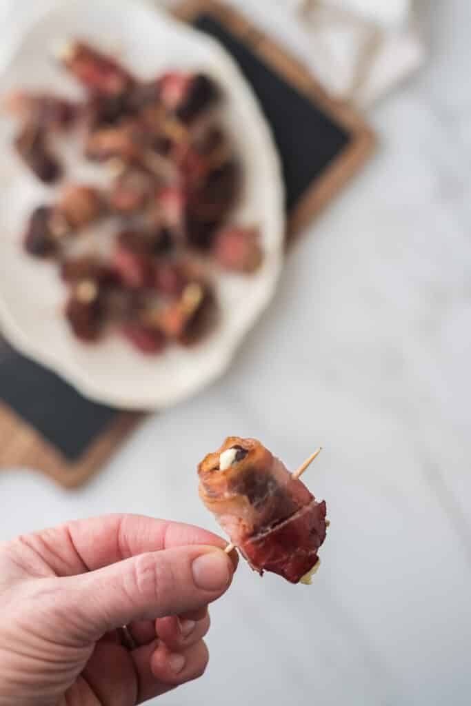 hand holding toothpick with bacon wrapped dates stuffed with goat cheese, platter with more behind