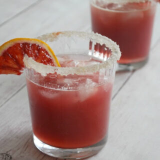 A white wooden table with glasses topped with sugar and blood orange slices, filled with blood orange bourbon smash cocktail 