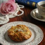A plate with an apple cheddar bacon scone and butter with tea, roses, and book in the background