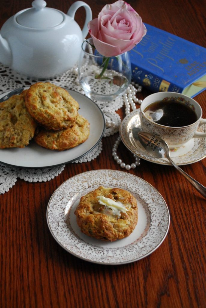 A cup of tea, teapot, rose, book, and two plates of apple cheddar bacon scones on a wooden table with pearls and doily