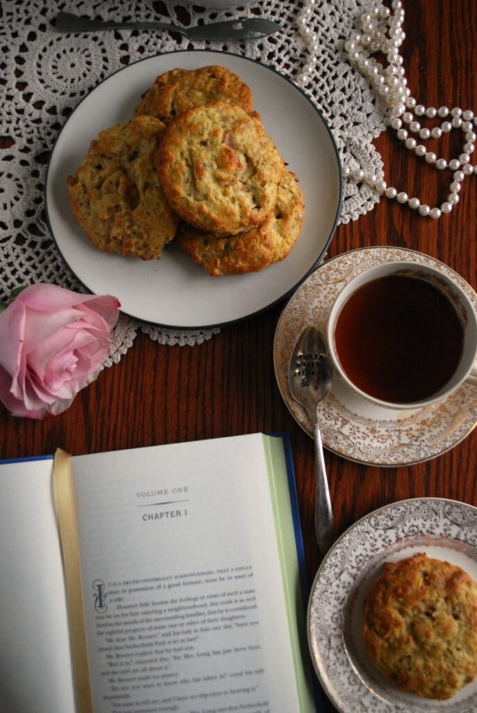 Two plates of apple bacon cheddar scones on a table with tea, a rose, books, a pearl necklace, and a white doily 