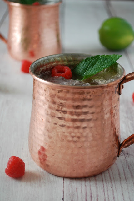 Two different Moscow Mule mugs on a white table with mint and raspberries