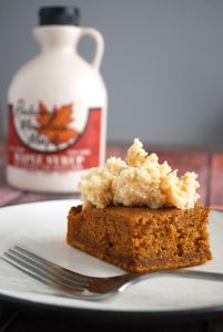 pumpkin gingerbread cake topped with frosting on plate with jug of maple syrup behind