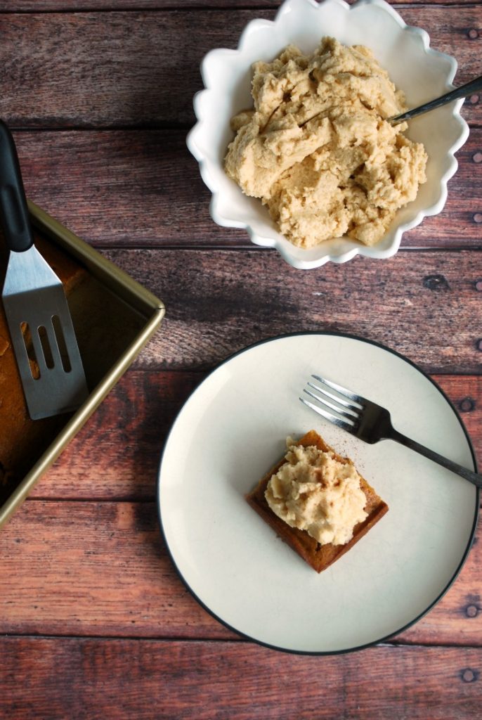 A white plate with a piece of pumpkin maple gingerbread cake, a cake pan with cake, and a bowl with Earl Grey frosting all on a wooden table