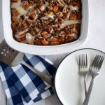 A white serving dish full of cinnamon roll breakfast casserole, on a white table with folding napkins, forks, and a stack of white plates