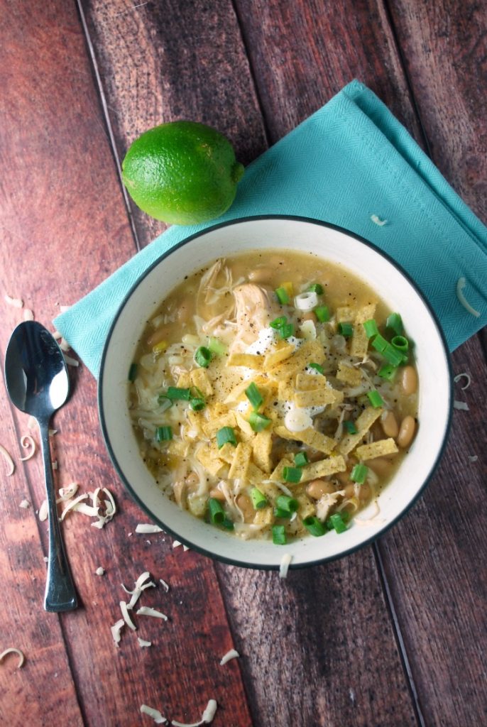 A wooden table topped with a bowl of white chicken chili, a spoon, I blew napkin, and a whole lime