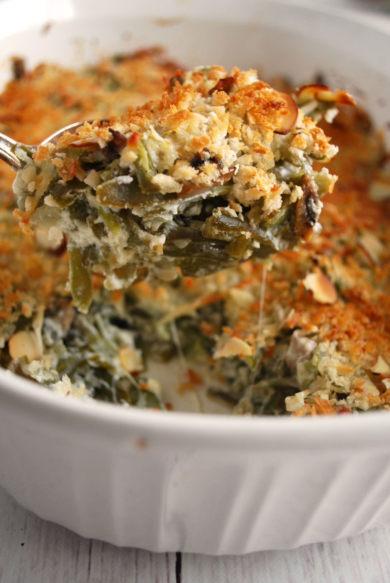 A spoon holding up a bite of green bean casserole over a white to serving dish of green bean casserole