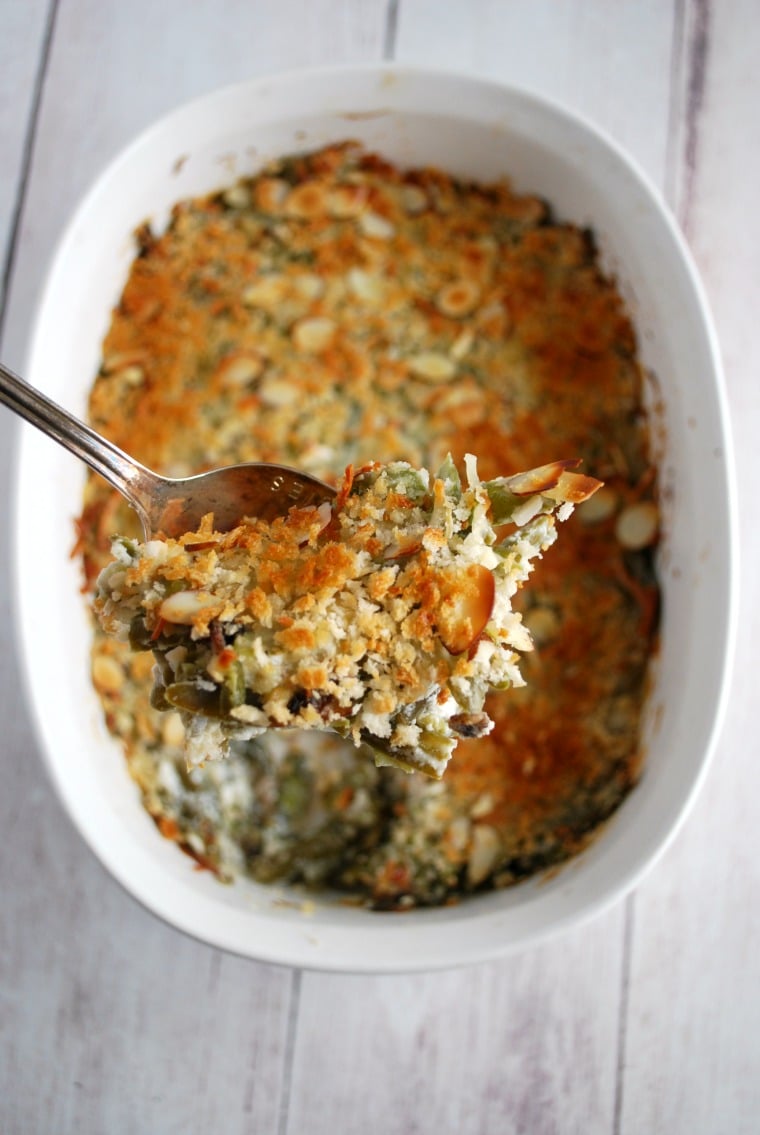 A spoon holding up a serving of green bean casserole over a white serving dish on a white wooden table