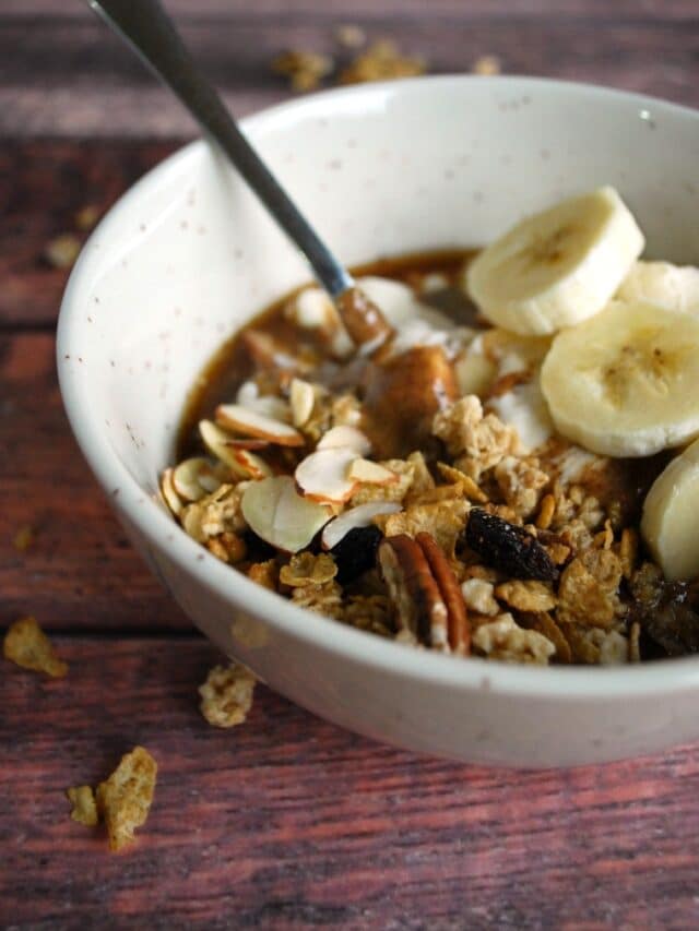 A white bowl of granola and yogurt parfait with banana slices and the silver spoon on a wooden table