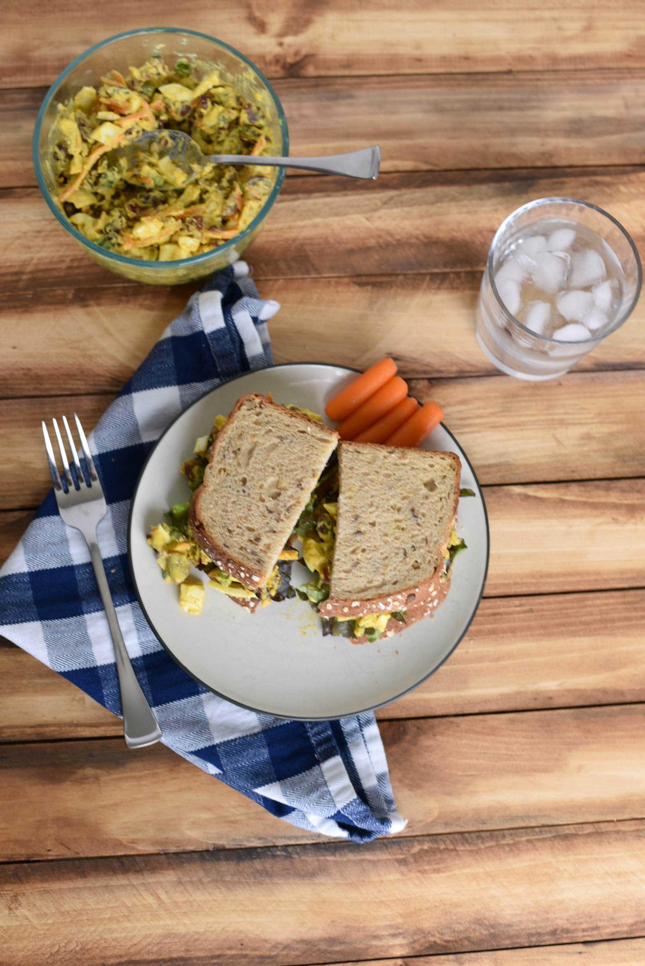 A wooden table with a bowl of curried egg salad, a plate with a sandwich and carrots, and a glass of water