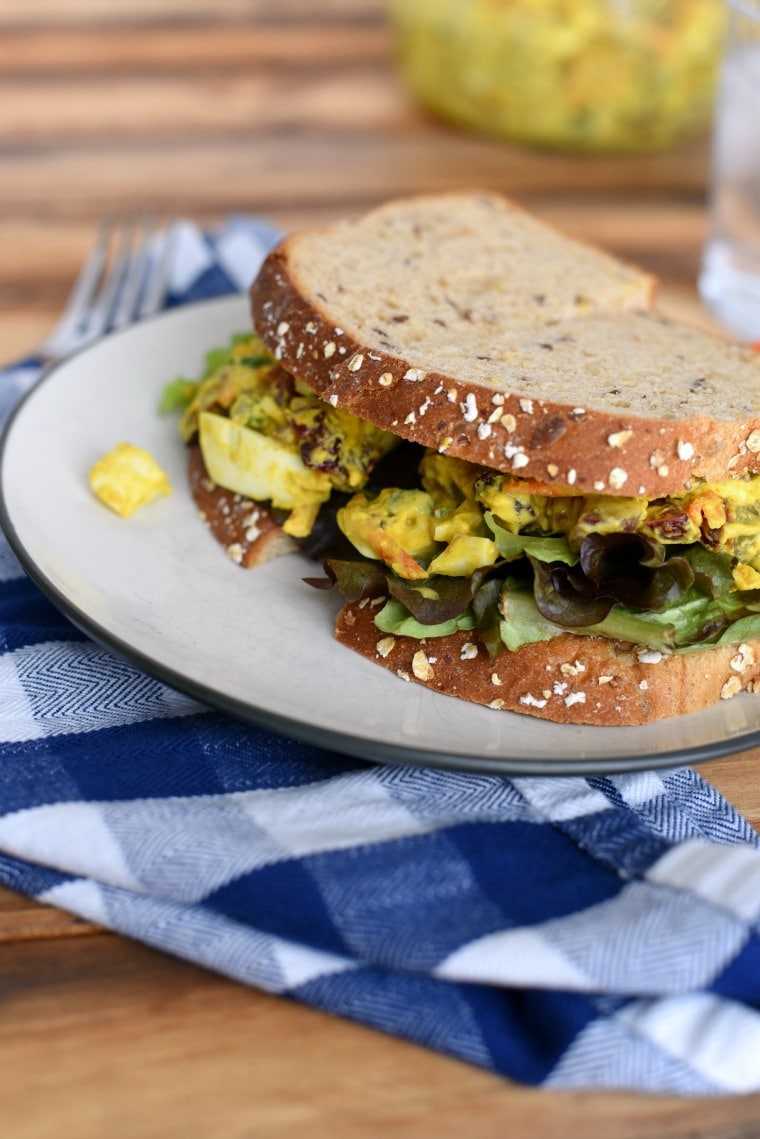 A white plate with a curried egg salad sandwich on a wooden table with a blue checked napkin