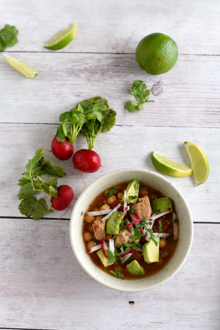 A white wooden table with a bowl of pork posole, radishes, and limes