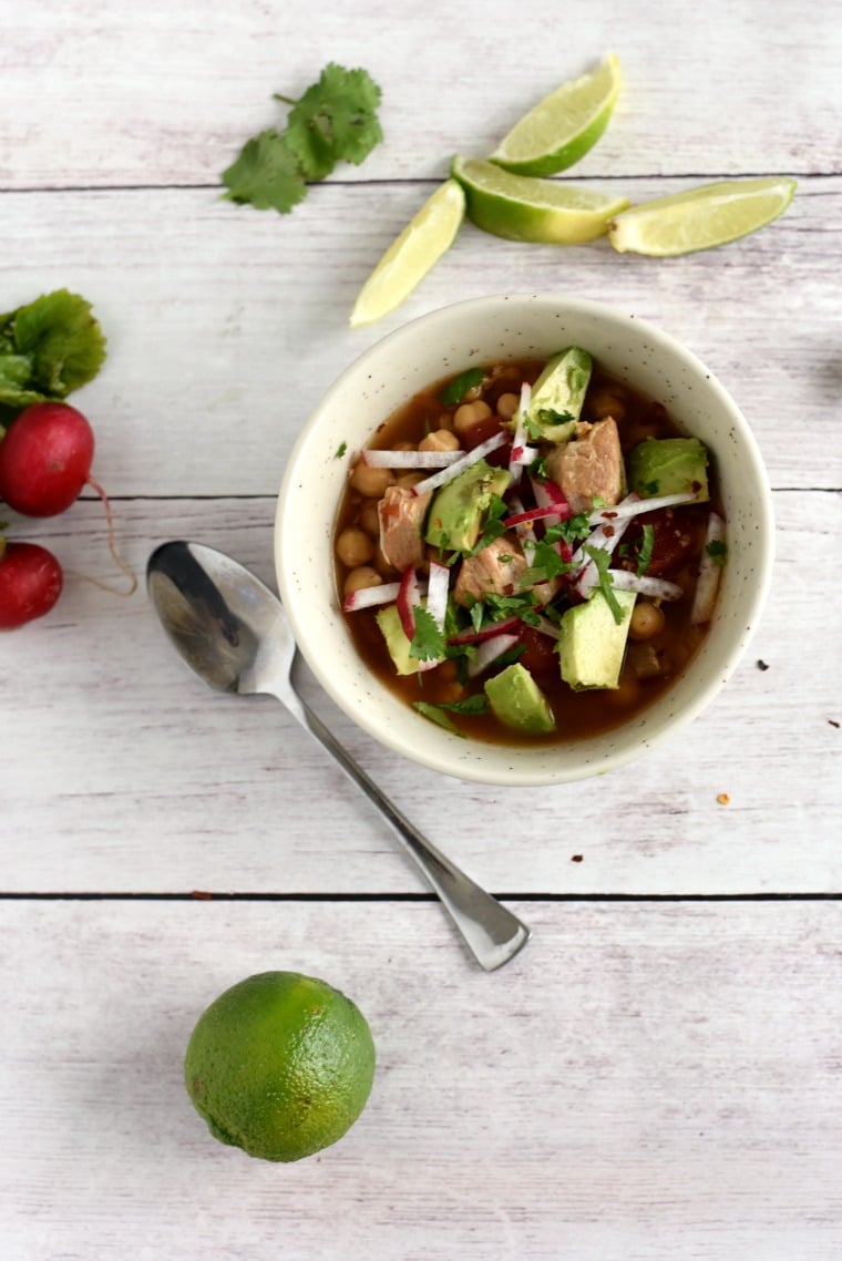 A white wooden table with a bowl of pork posole, a spoon, radishes, and limes