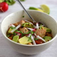 A white bowl of pork posole with radishes and limes in the background