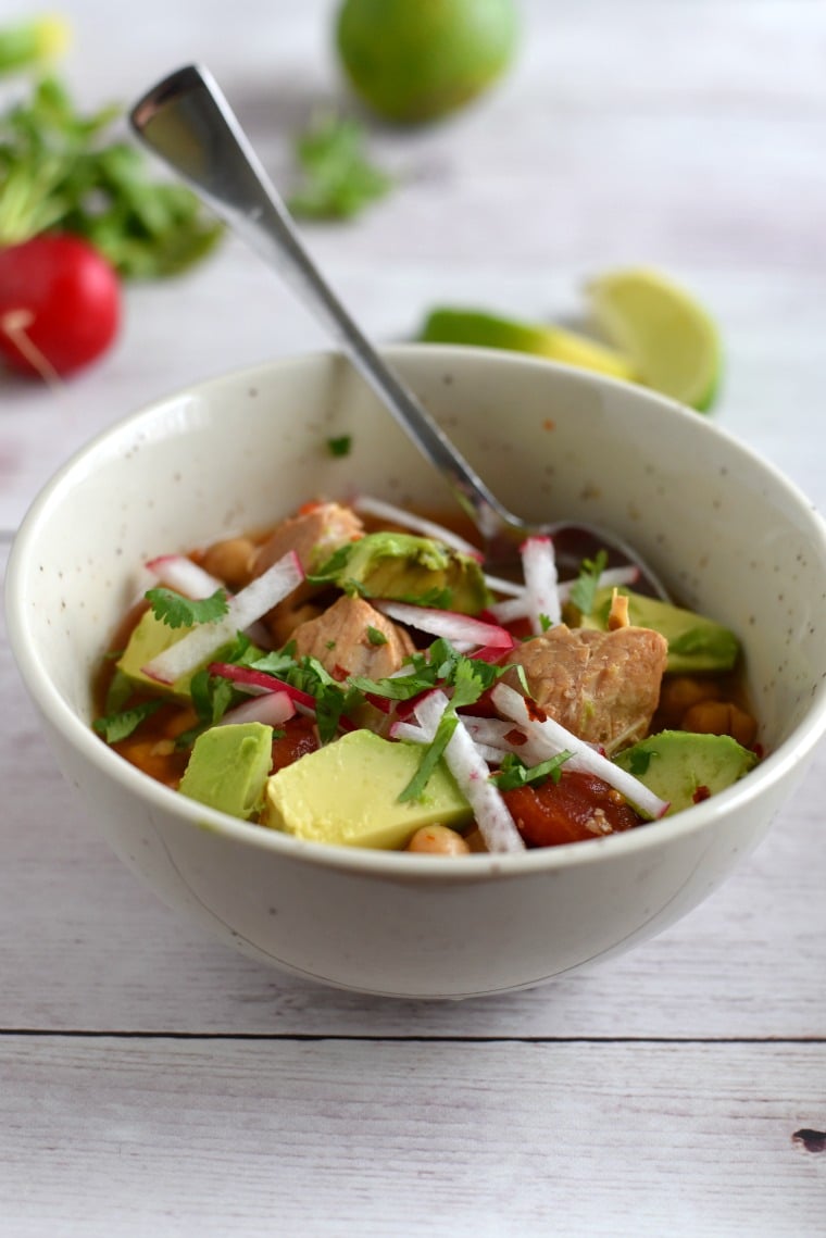 A white bowl of pork posole with radishes and limes in the background 