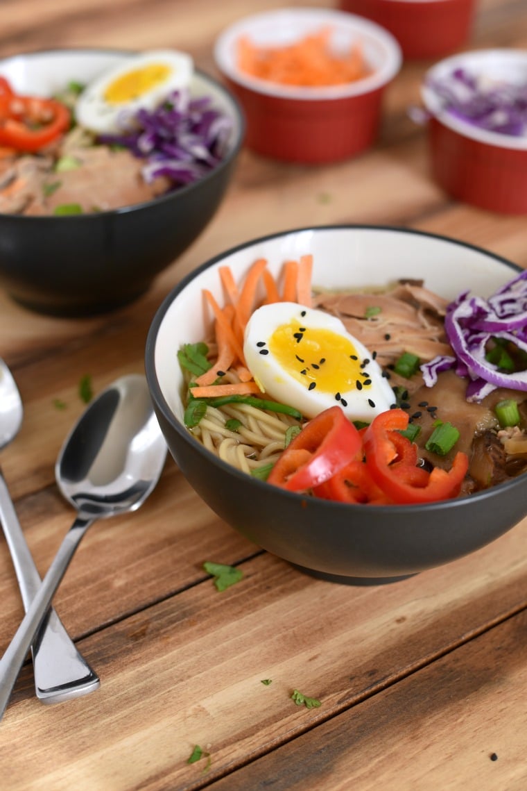 Two bowls of crock pot chicken ramen with hard boiled eggs and two spoons on a wooden table