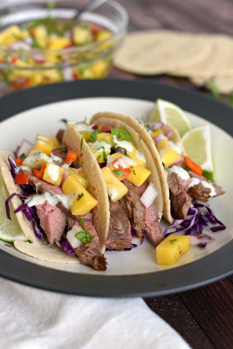 A white and black plate with three flank steak tacos and a bowl of mango salsa in the background 