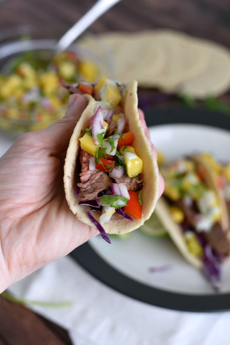 Hand holding flank steak taco with pineapple mango salsa, cabbage, and cilantro lime sauce
