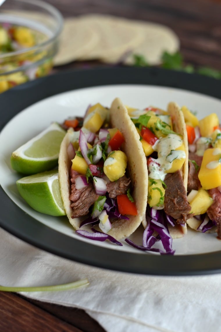 A plate with three mango flank steak tacos and lime wedges