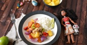 A plate of Hawaiian chicken and veggies on a wooden table with a Moana doll