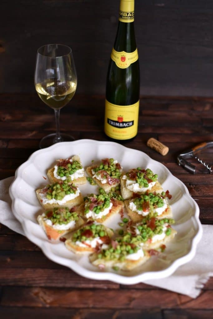 A bottle and glass of sparkling wine with a platter of ricotta pea toasts on a wooden table