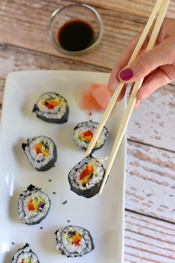 A woman with magenta nail polish picking up a piece of rainbow veggie sushi with chopsticks 