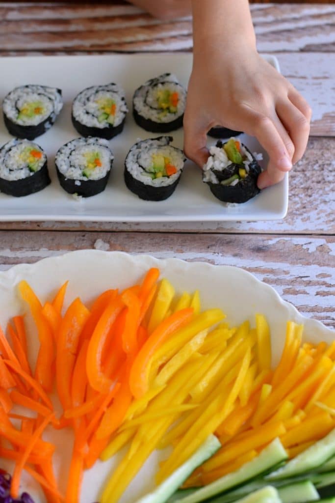 A child picking up a piece of rainbow veggies sushi from a plate of sushi and a separate plate of sliced veggies 