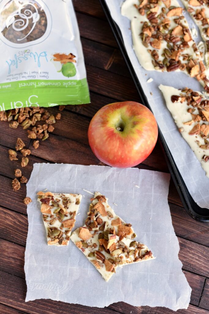 A wooden table with a spilled bag of granola, an apple, and pieces of apple cinnamon white chocolate bark on pieces of parchment paper