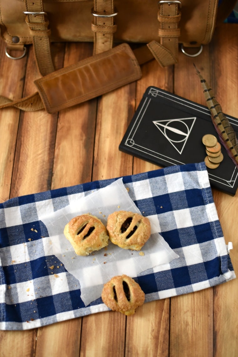 A wooden table with a leather bag, a white and blue checked napkin, cauldron cakes, a feather, coins, and a book with the symbol of the Deathly Hallows 