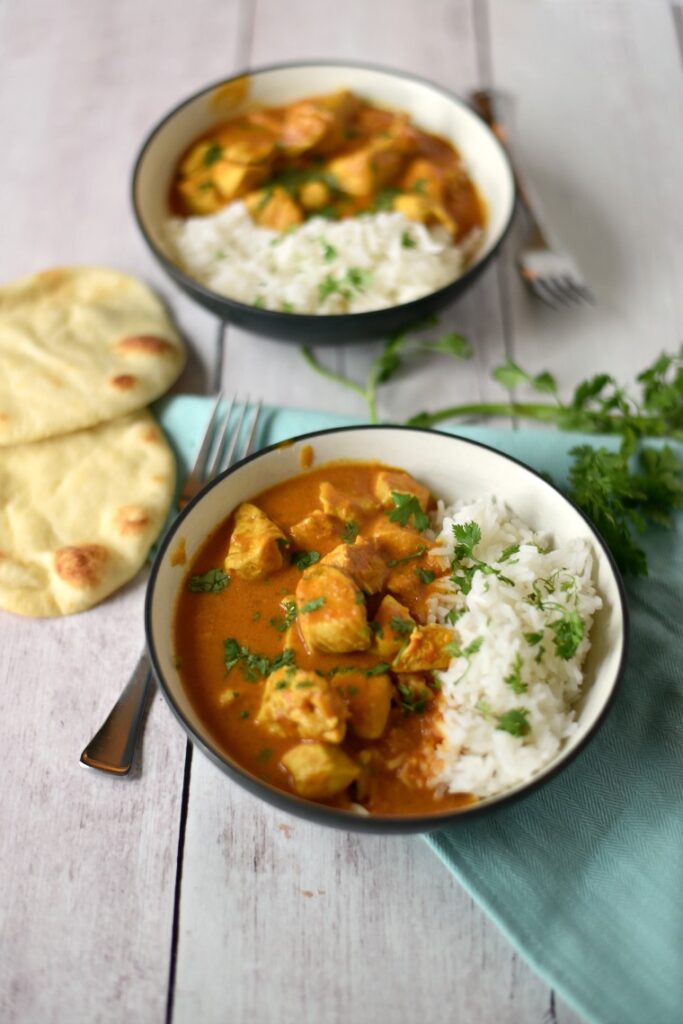 A white wooden table with two bowls of chicken tikka masala and rice, with naan on the side