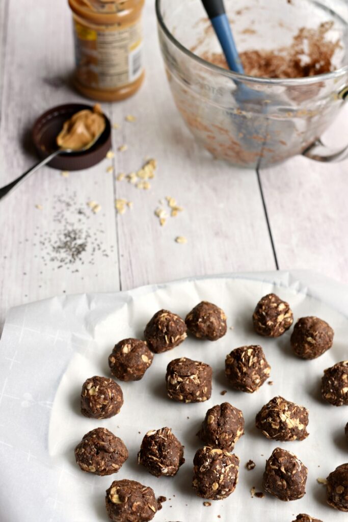 A plate of chocolate peanut butter energy balls on a white wooden table with peanut butter and a mixing bowl