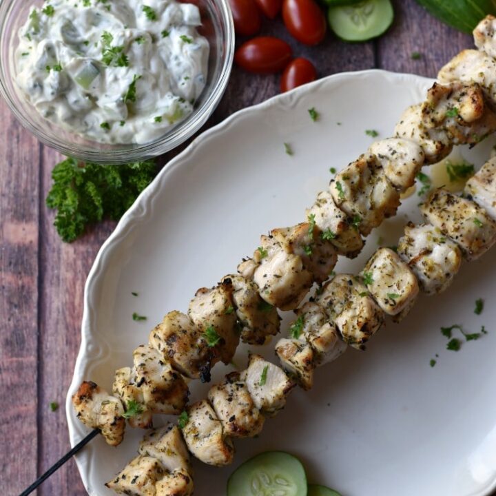 grilled chicken skewers on white platter with tzatziki, cucumbers and tomatoes