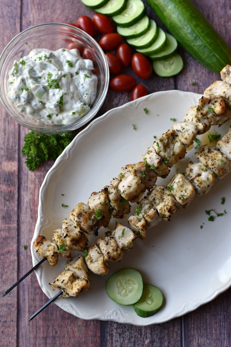 grilled chicken skewers on white platter with tzatziki, cucumbers and tomatoes